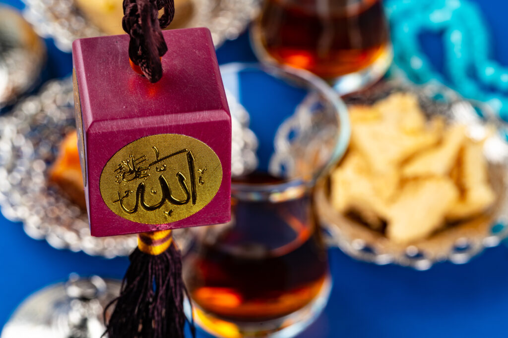 Bordeauxrode islamitische amulet met het woord 'Allah' in gouden Arabische kalligrafie, met op de achtergrond een glas Turkse thee en traditionele zoetigheden – symbool voor gastvrijheid in halalvriendelijke vakanties.