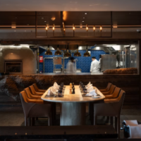 Meast Steakhouse heeft een sfeervol interieur met warme verlichting en een open keuken. Donker steakhouse-interieur met lange gedekte tafel en open keuken op AROYA